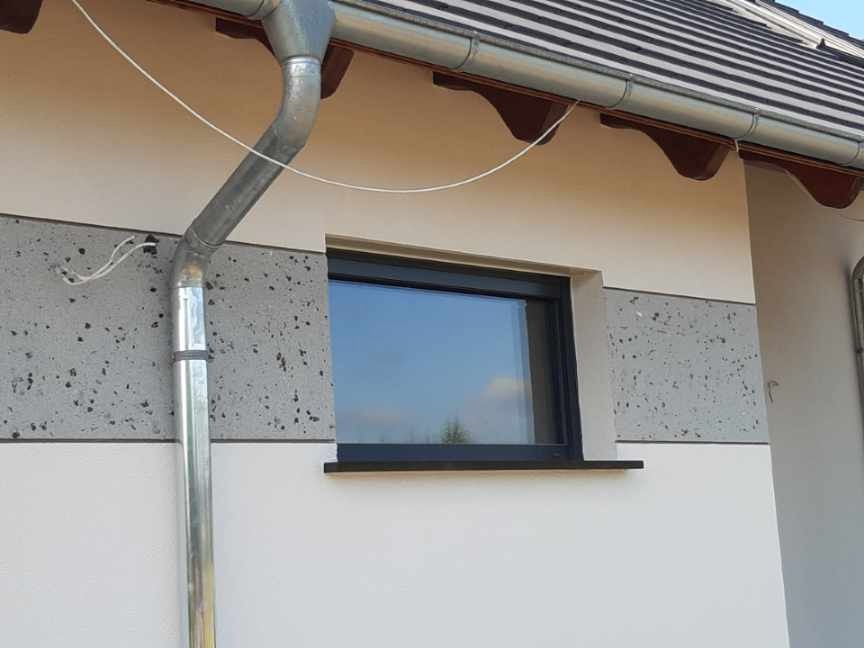 Fragment elewacji domu z oknem i rynną, widoczny pas szarego tynku z ciemnymi plamkami, kontrastujący z gładką, jasną powierzchnią ściany.