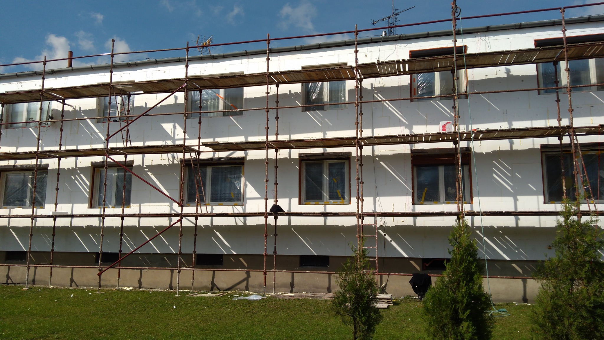 Budynek wielorodzinny w trakcie prac izolacyjnych elewacji, widoczne rusztowania, zabezpieczone okna folią i częściowo przyklejone płyty izolacyjne, w tle błękitne niebo z chmurami.