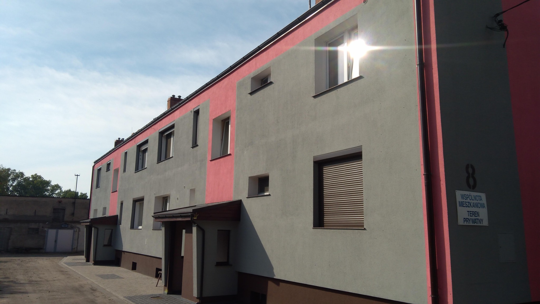Ocieplony budynek wielorodzinny z szarą elewacją i różowymi akcentami, widoczne okna z roletami, tabliczka informacyjna 'Wspólnota Mieszkaniowa Teren Prywatny'.
