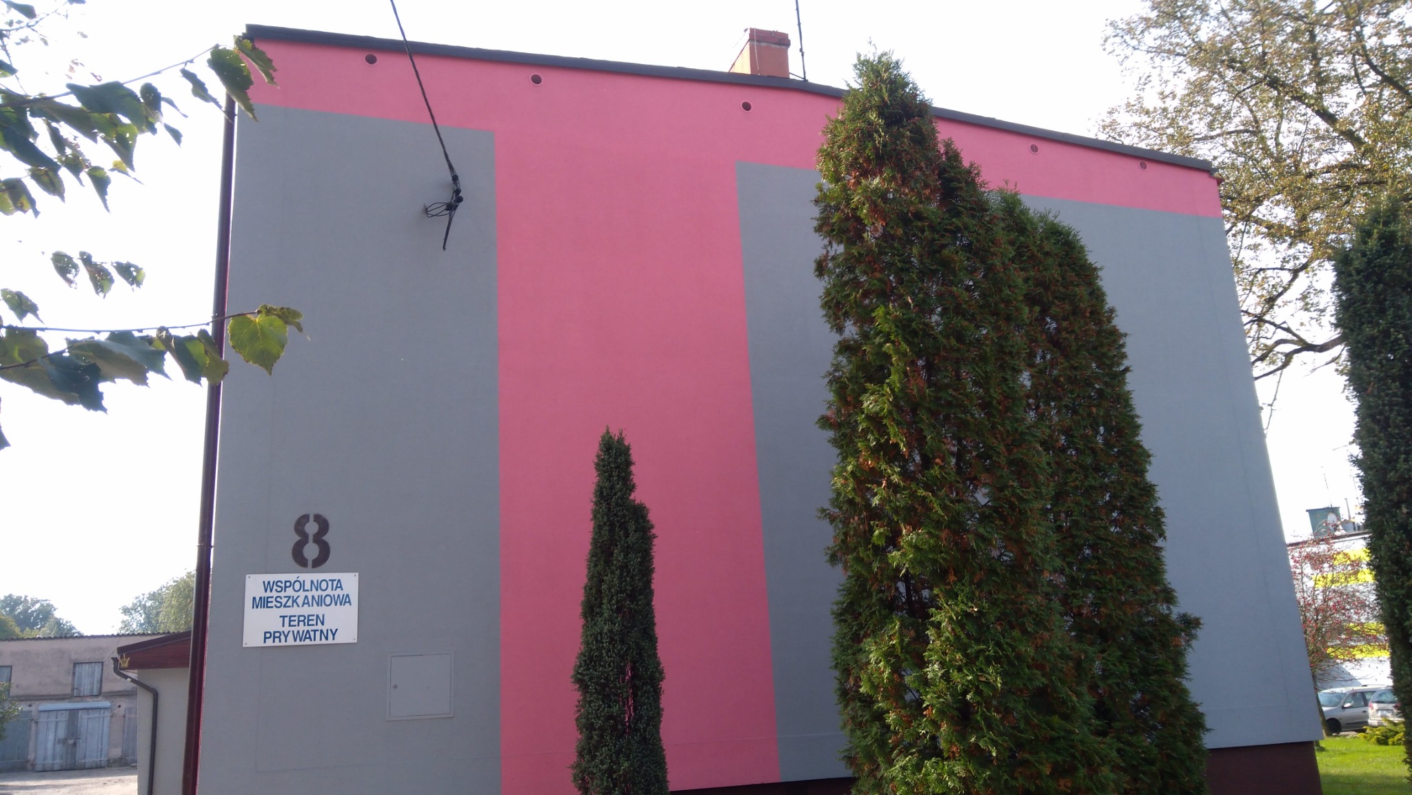 Dwukolorowa elewacja budynku wielorodzinnego w odcieniach szarości i różu, z widoczną tabliczką informacyjną 'Wspólnota Mieszkaniowa' oraz trzema iglakami w różnej wielkości.