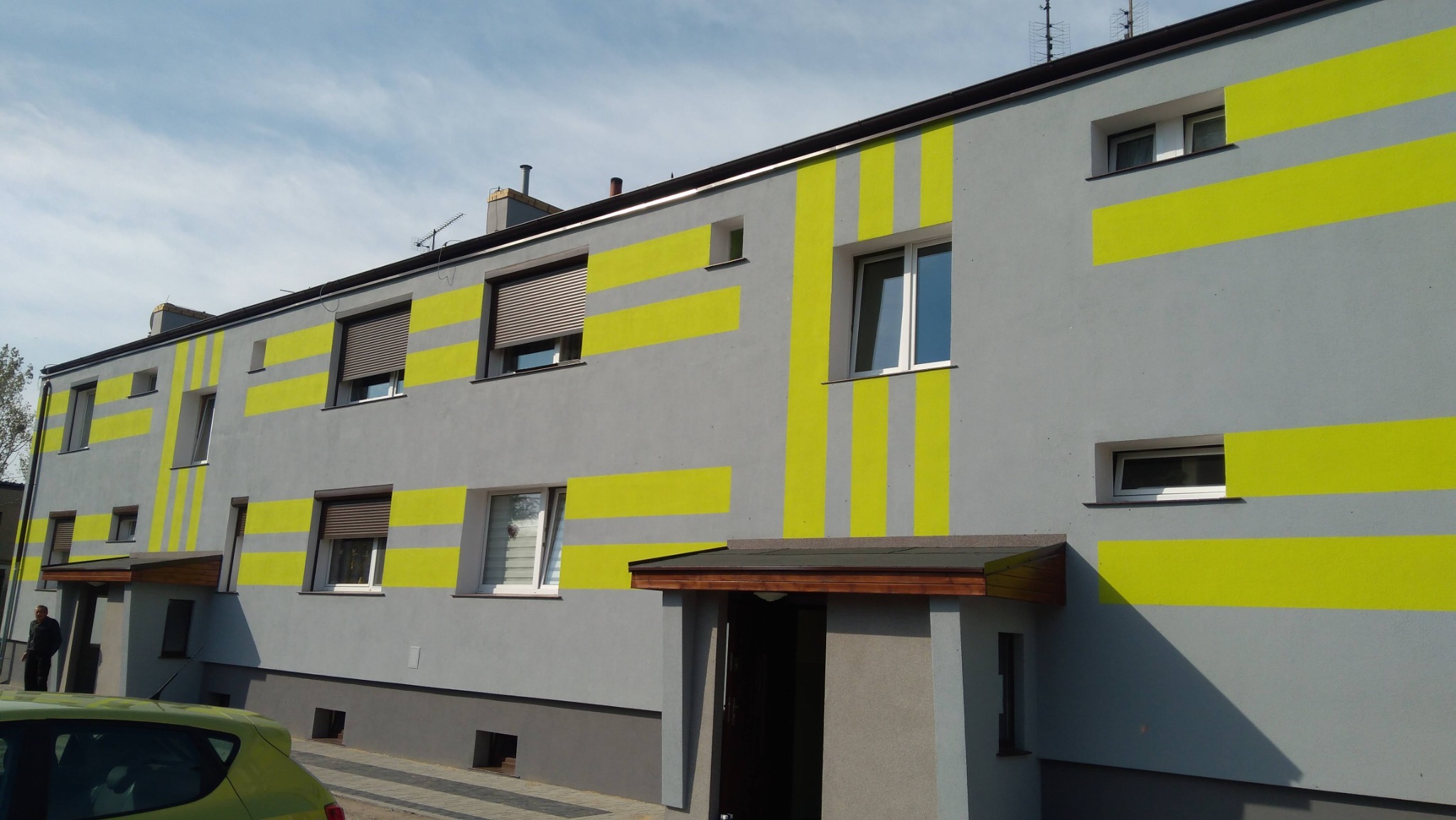 Szary budynek wielorodzinny z jaskrawożółtymi pasami na elewacji, widoczne okna z roletami i bez, oraz wejście z drewnianym zadaszeniem.