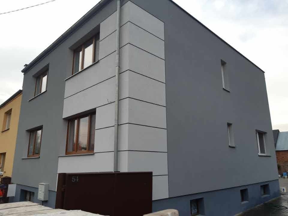 Dwukondygnacyjny dom po remoncie elewacji, z szarymi ścianami i jasnoszarymi poziomymi pasami dekoracyjnymi na narożniku. Widoczny numer domu '54' nad brązową bramą wjazdową.