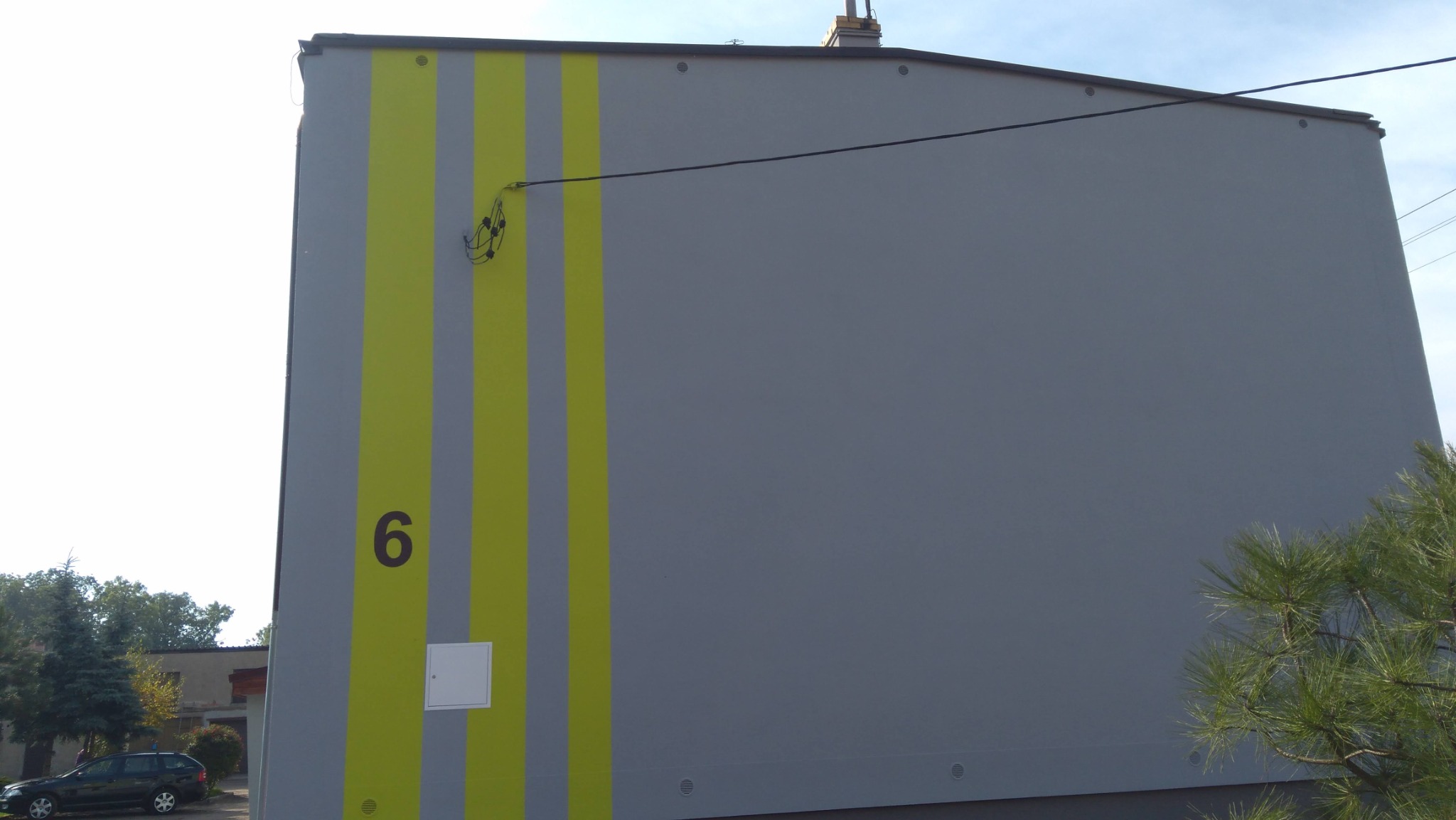 Szara ściana budynku z pionowymi, żółtymi pasami, numerem '6' i skrzynką elektryczną, widoczne drzewa i samochód w tle.