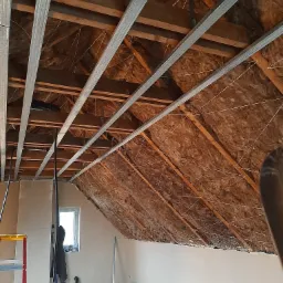 Poddasze z ociepleniem wełną mineralną, widoczne metalowe profile sufitowe w trakcie montażu, fragment ściany po szpachlowaniu, okno i drabina w tle.