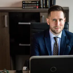 Portret mężczyzny w garniturze siedzącego za biurkiem z laptopem, w tle regał z książkami i dokumentami.