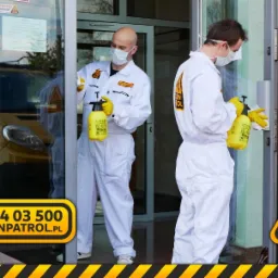Dw&oacute;ch mężczyzn w białych kombinezonach i maskach dezynfekuje wejście do budynku, używając ż&oacute;łtych spryskiwaczy. W tle widoczny ż&oacute;łty samoch&oacute;d.