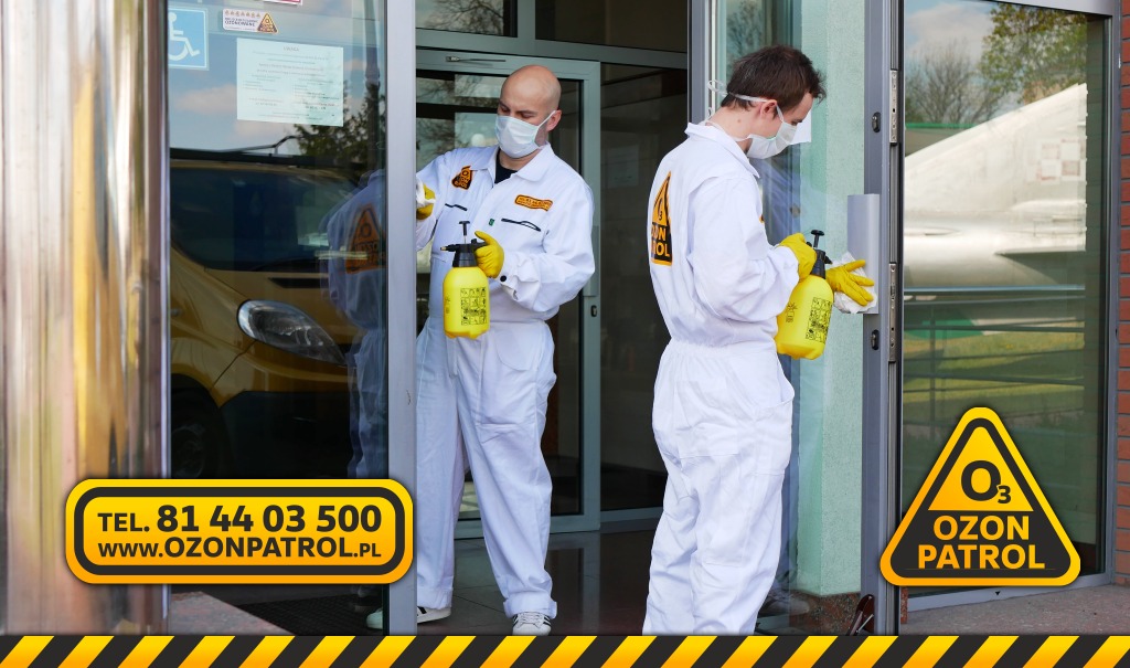 Dwóch mężczyzn w białych kombinezonach i maskach dezynfekuje wejście do budynku, używając żółtych spryskiwaczy. W tle widoczny żółty samochód.