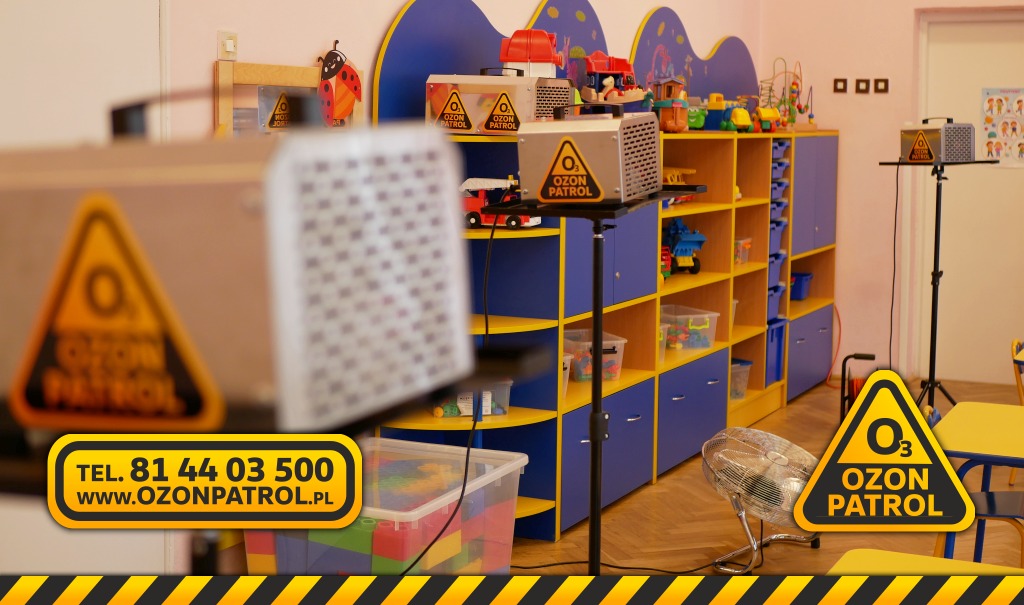 Ozonowanie przedszkola: kilka urządzeń Ozon Patrol na statywach w kolorowym pomieszczeniu z półkami pełnymi zabawek, wentylator na podłodze, w tle dekoracje ścienne z motywem biedronki.