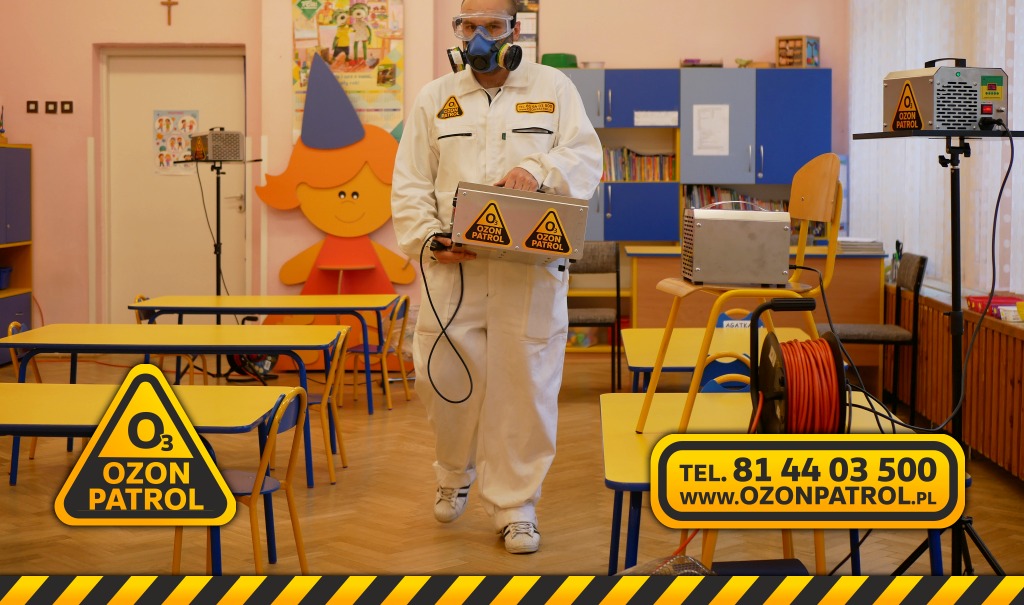 Pracownik w kombinezonie i masce ochronnej niesie urządzenie do ozonowania w sali przedszkolnej z żółtymi stolikami i krzesłami; w tle widoczne dekoracje ścienne i inne urządzenia do ozonowania.