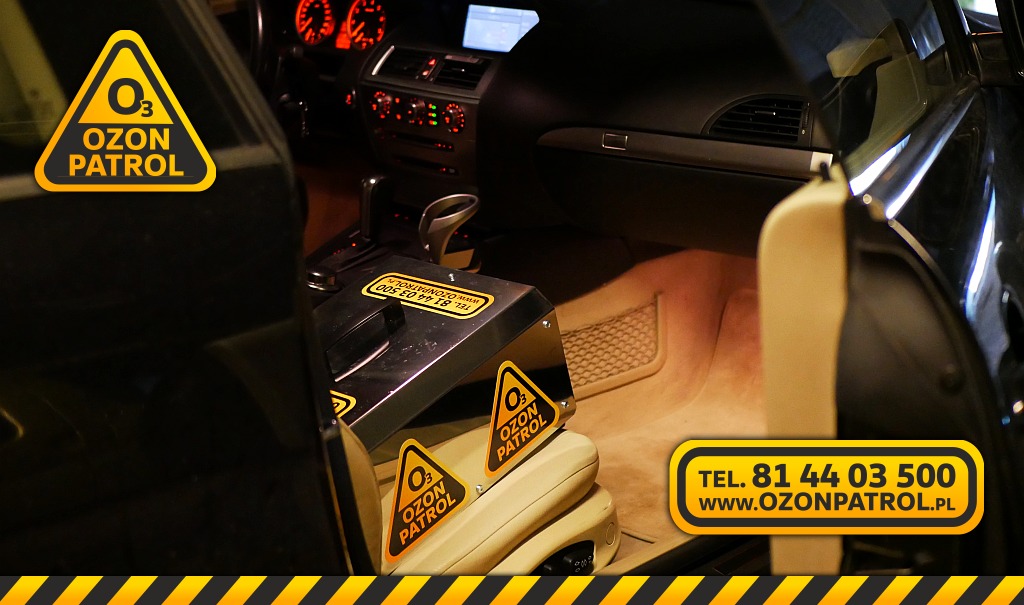 Ozonator z naklejkami Ozon Patrol umieszczony wewnątrz samochodu podczas procesu odkażania wnętrza, widoczne elementy deski rozdzielczej i beżowej tapicerki.
