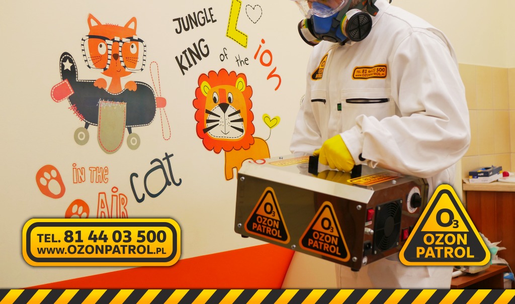 Osoba w kombinezonie ochronnym i masce obsługuje ozonator powietrza w pomieszczeniu z dziecięcymi malunkami na ścianie, widoczne logo Ozon Patrol.