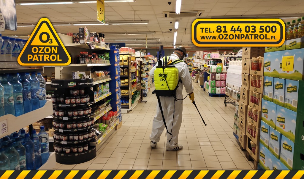 Pracownik w kombinezonie ochronnym i masce rozpyla środek dezynfekujący w supermarkecie, używając opryskiwacza plecakowego Titan. Widoczne regały z produktami spożywczymi i naklejki reklamowe Ozon...