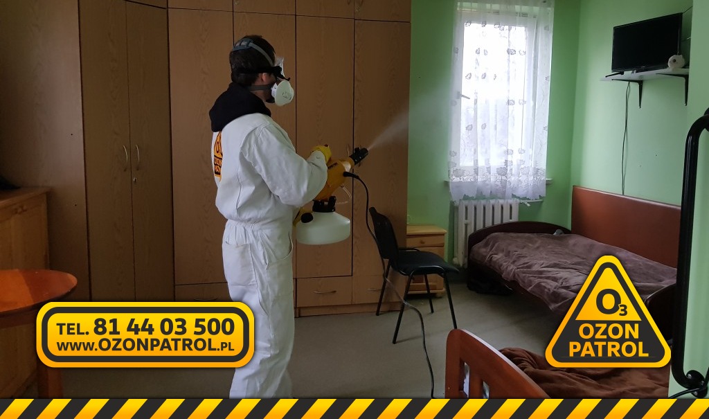 Osoba w kombinezonie ochronnym i masce przeprowadza ozonowanie pomieszczenia za pomocą profesjonalnego generatora ozonu. Widoczny sprzęt do zamgławiania ULV w pokoju z łóżkiem i szafą.