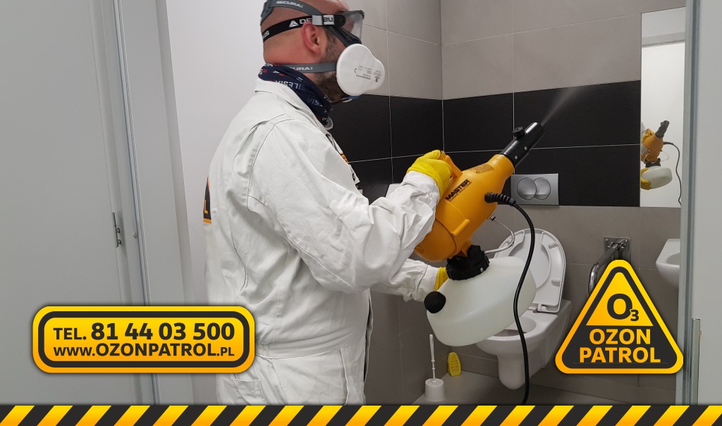 Pracownik w kombinezonie ochronnym, masce i okularach ochronnych, używa żółtego zamgławiacza Master do dezynfekcji toalety. Widoczny napis Ozon Patrol na kombinezonie i logo firmy w rogu zdjęcia.