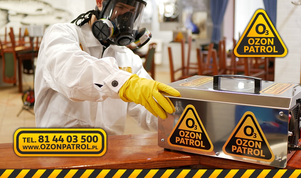 Osoba w masce ochronnej i żółtych rękawicach obsługuje ozonator Ozon Patrol w pomieszczeniu, widoczne ostrzegawcze naklejki z logo firmy na urządzeniu.