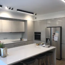 Nowoczesna kuchnia z szarymi, lakierowanymi frontami szafek, oświetleniem szynowym i wyspą z blatem imitującym marmur, uzupełniona o drewniane hokery