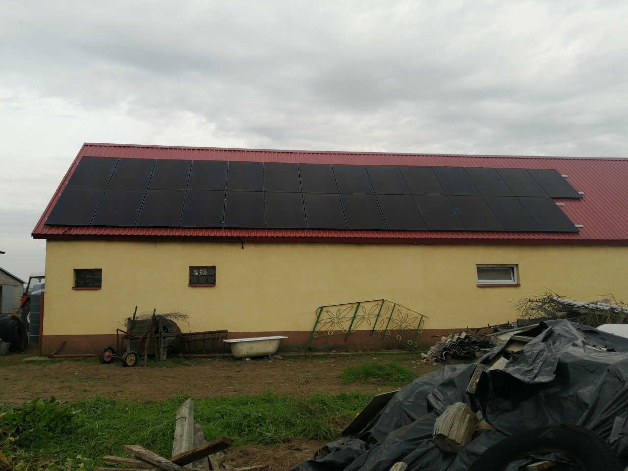 Żółty budynek gospodarczy z czerwonym dachem pokrytym panelami słonecznymi; w tle pochmurne niebo.