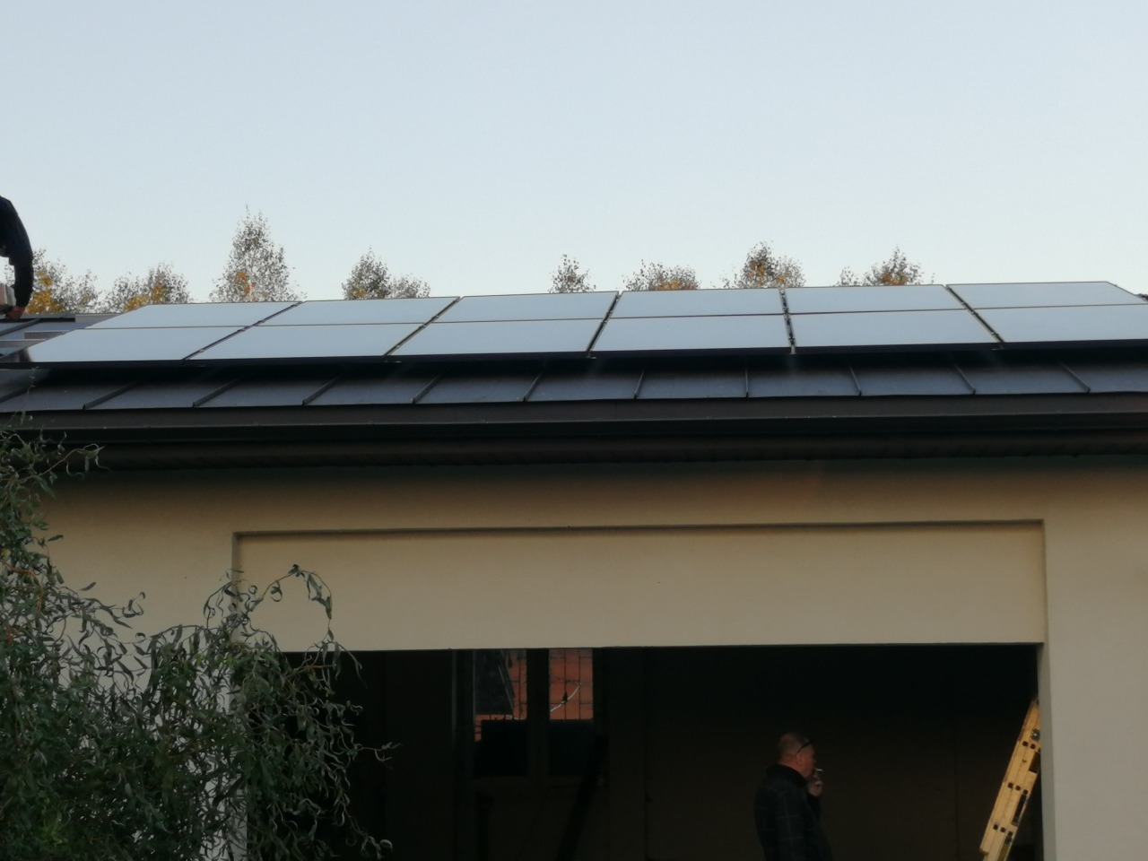 Instalacja paneli słonecznych na dachu budynku garażowego, widoczny fragment osoby montującej i mężczyzna w tle obok drabiny.