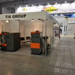 Ekspozycja firmy TIS Group na targach, prezentująca dwa kotły: jeden z palnikiem retortowym i podajnikiem, drugi segmentowy z pomarańczowymi drzwiczkami, na tle stoiska i innych ekspozycji.