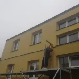 Pracownik na rusztowaniu mocuje metalową listwę na żółtej elewacji budynku, widoczne okna i pochmurne niebo.