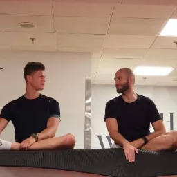 Dwóch mężczyzn siedzi po turecku na czarnym, lekko wygiętym podeście w jasnym pomieszczeniu z białymi ścianami i sufitem kasetonowym, patrząc na siebie.