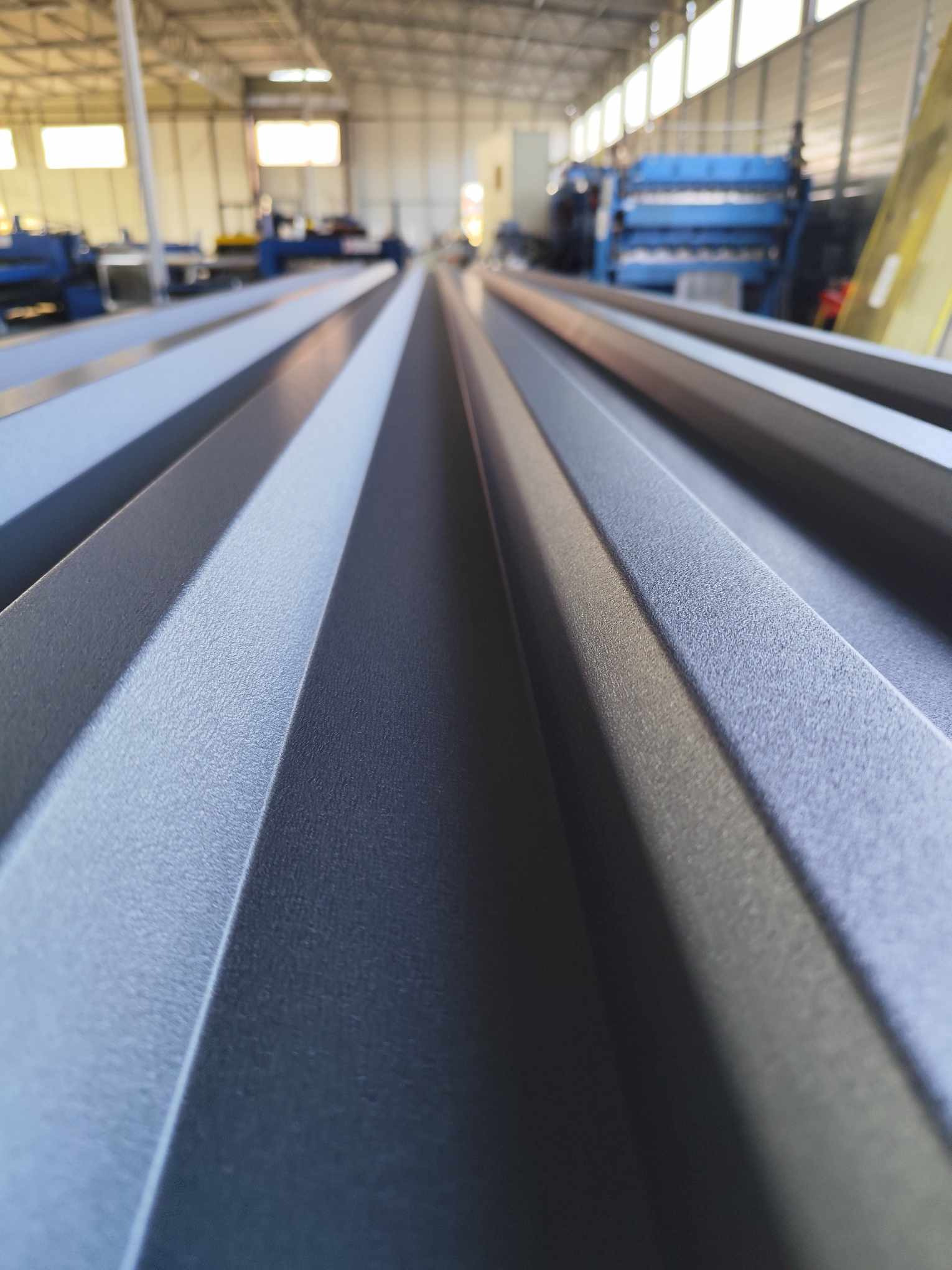 Szary arkusz profilowanej blachy, widok z bliska, z maszyną do obróbki blach w tle hali produkcyjnej.