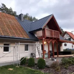 Dom jednorodzinny z częściowo zdjętym i częściowo nowym, grafitowym pokryciem dachowym, widoczne rusztowania i drabina, balkon z drewnianą konstrukcją.