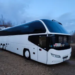 Biały autokar turystyczny marki Progress zaparkowany na żwirowym placu, widoczny z boku, z budynkiem szkoły jazdy w tle i niebieskim niebem nad nim.