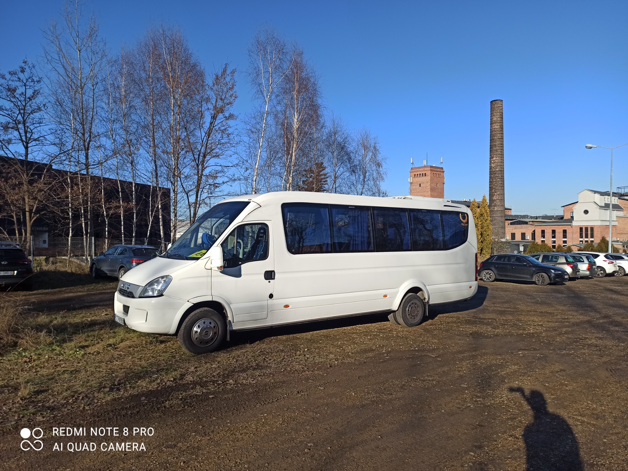 Biały bus pasażerski marki Iveco na tle ceglanego budynku z kominem w słoneczny dzień.