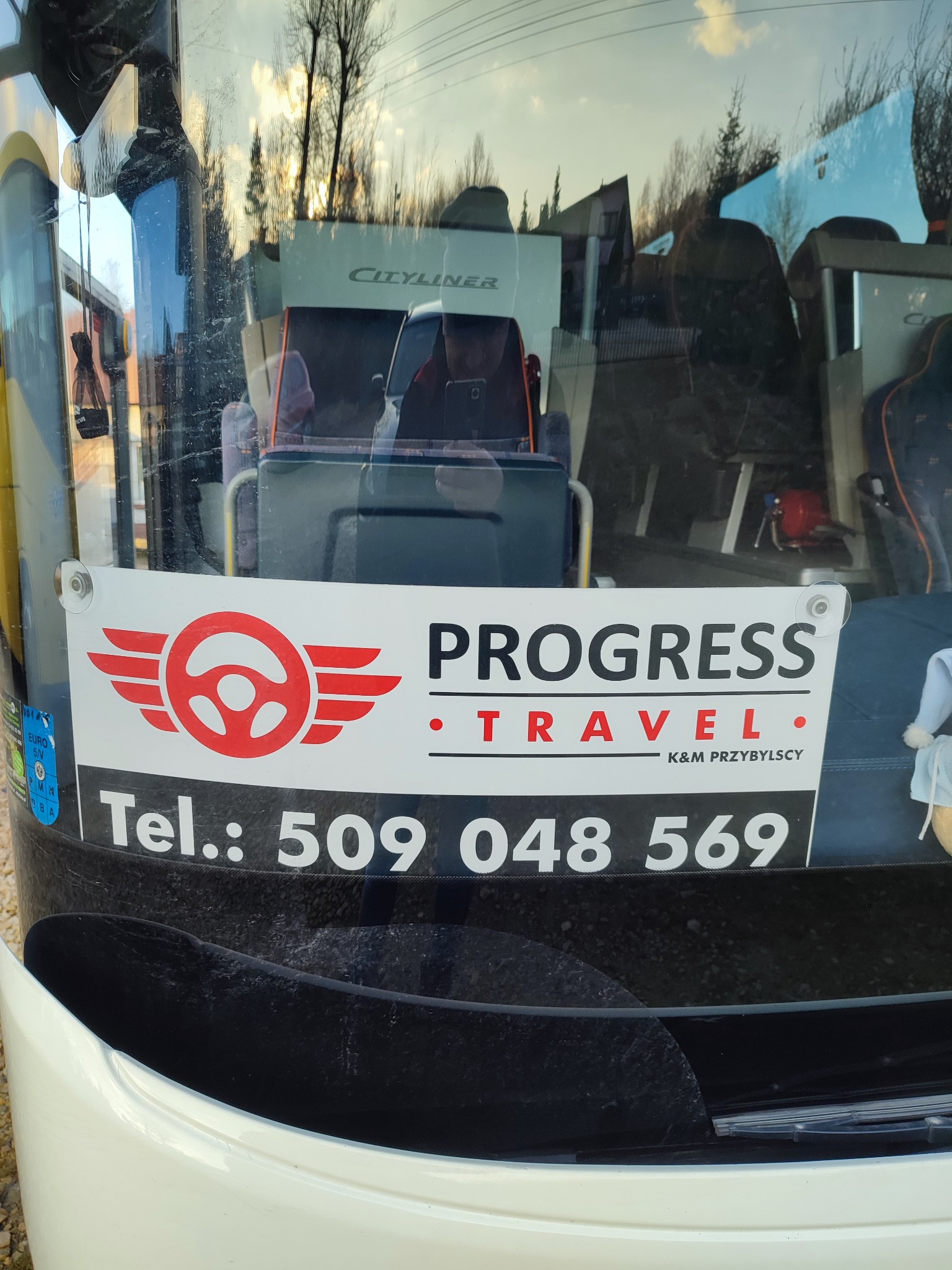 Reklama firmy Progress Travel z numerem telefonu 509 048 569 umieszczona na przedniej szybie autobusu Cityliner z widocznym wnętrzem pojazdu i odbiciem otoczenia.