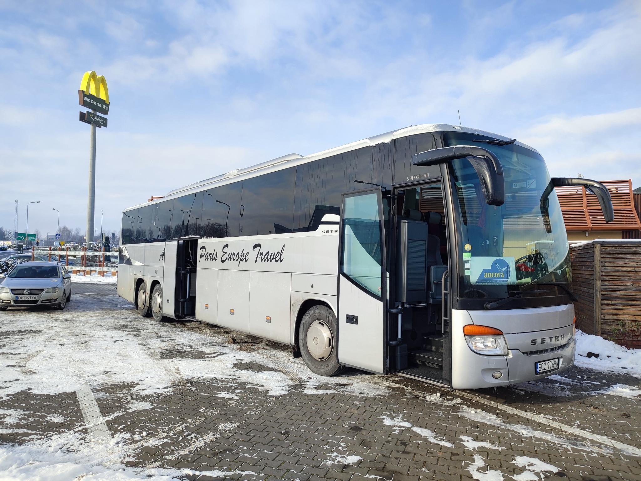 Srebrny autokar turystyczny Setra z otwartymi drzwiami na parkingu pokrytym śniegiem, z widocznym logo 'Paris Europe Travel', zaparkowany obok znaku McDonald's.