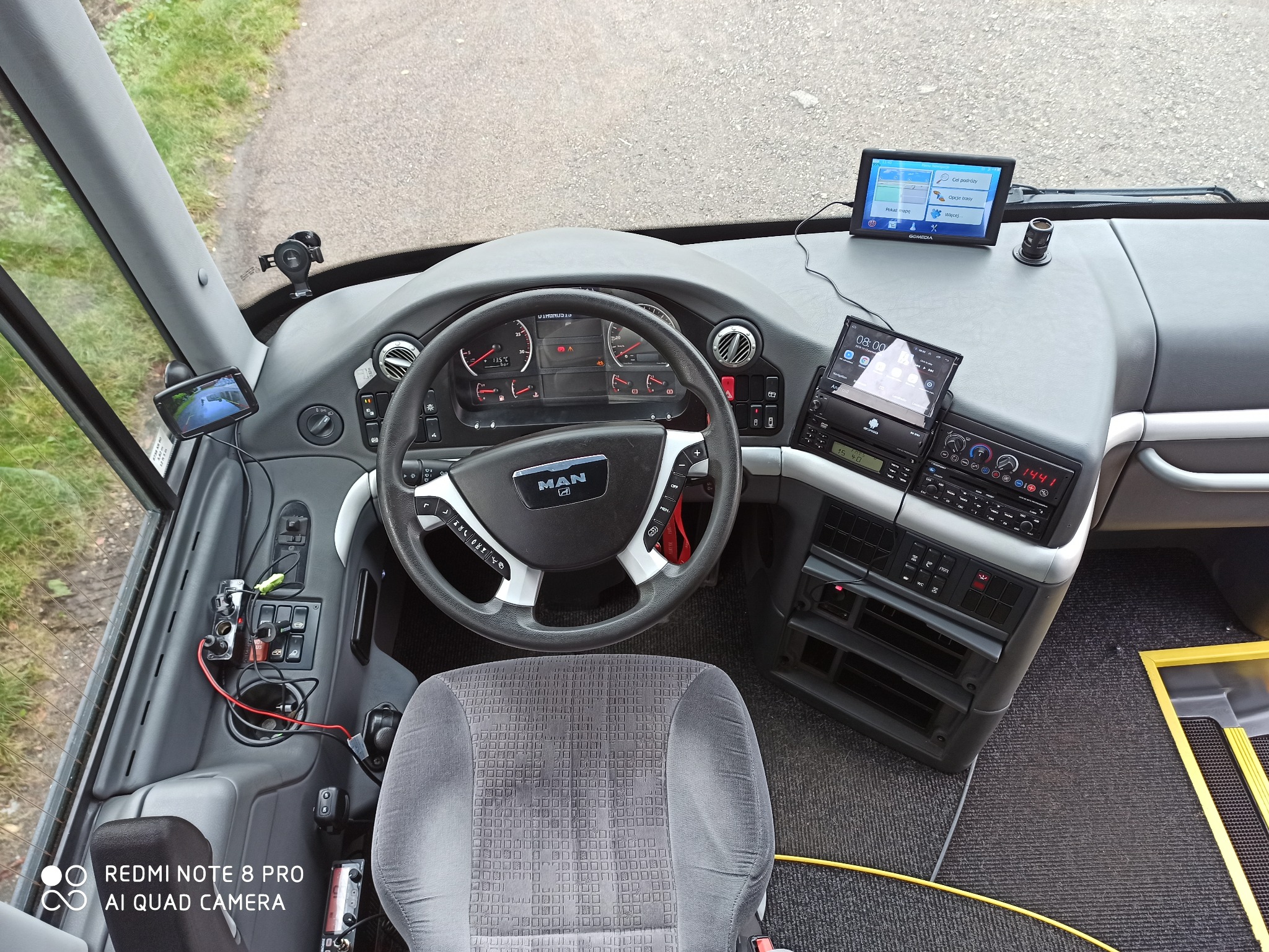 Wnętrze autobusu MAN z perspektywy kierowcy, widoczna deska rozdzielcza z nawigacją GPS, panelem sterowania, kierownicą i fotelem kierowcy, na przedniej szybie zamontowany dodatkowy monitor