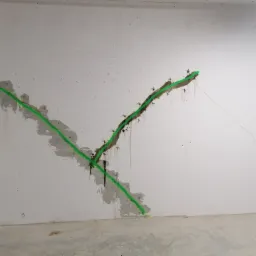 Betonowa ściana z zaznaczonym pęknięciem wypełnionym szarym materiałem i obrysowanym zieloną farbą, widoczne ślady wilgoci i metalowe kotwy w szczelinie.