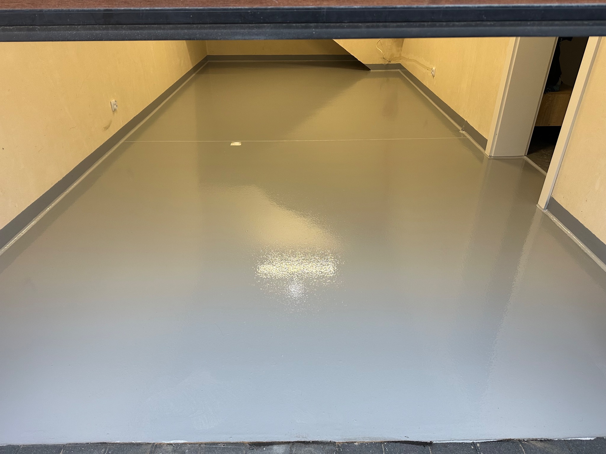 Szara, błyszcząca posadzka żywiczna w pomieszczeniu z beżowymi ścianami i szarymi listwami przypodłogowymi. Widok z góry, z lekkim refleksem światła na powierzchni.