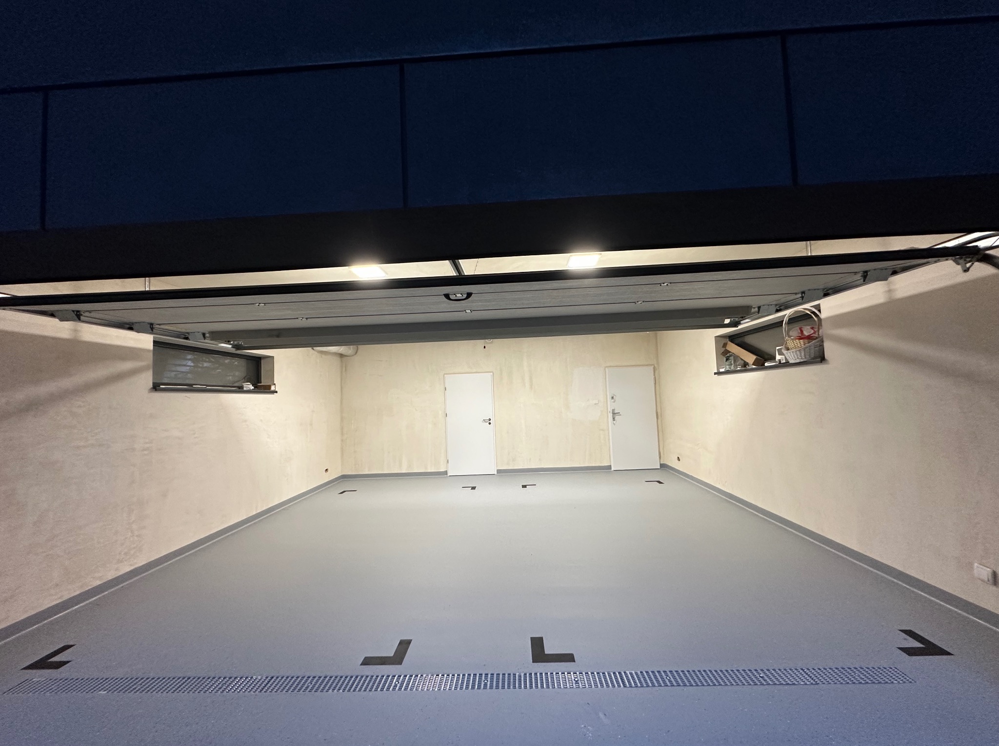 Szara posadzka żywiczna w garażu z odpływem liniowym i czarnymi znakami kierunkowymi, widok na dwie białe drzwi i okno z koszem.