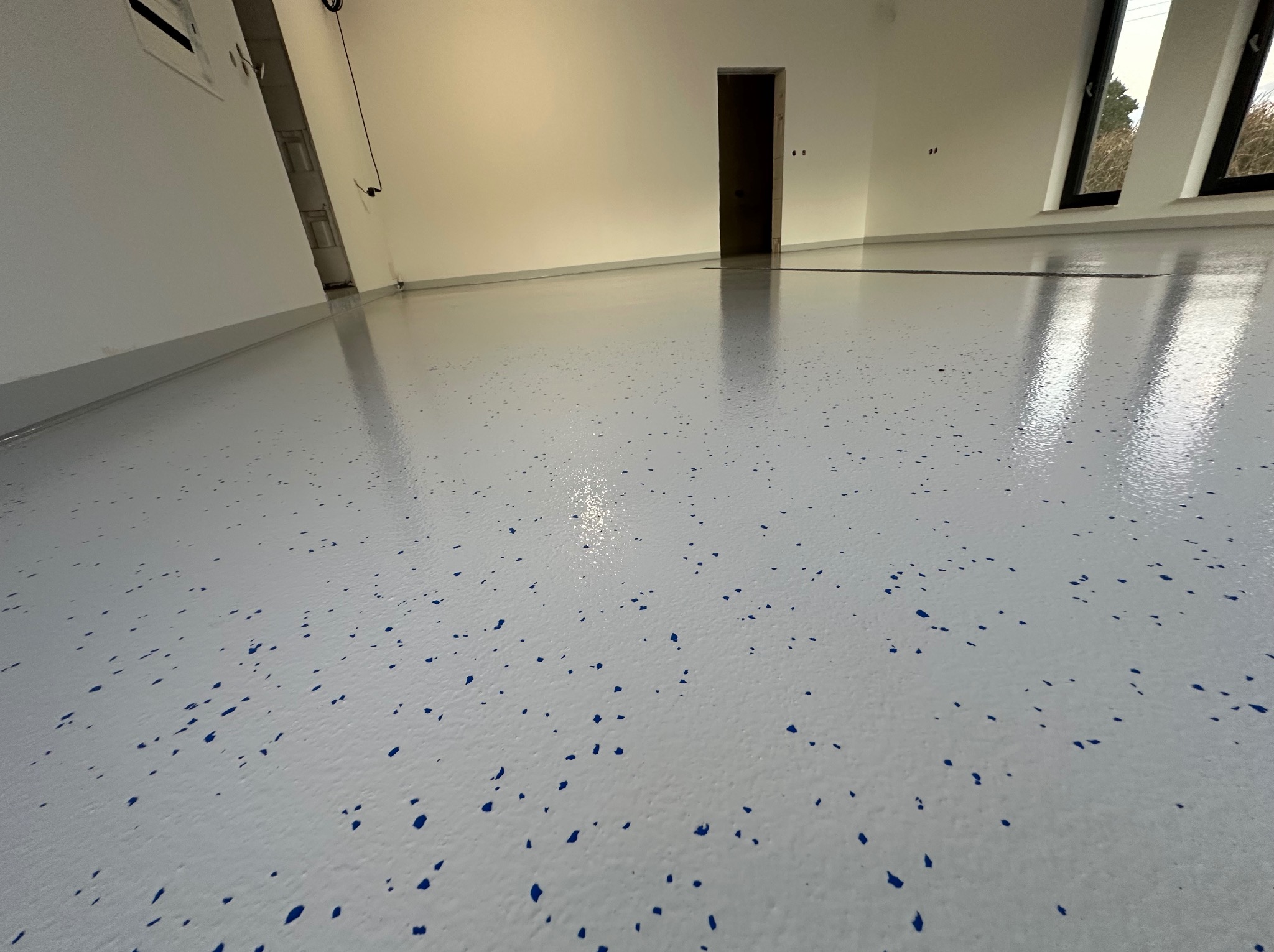 Biała posadzka żywiczna z niebieskimi elementami dekoracyjnymi w jasnym pomieszczeniu z oknami i drzwiami. Błyszcząca powierzchnia, świeżo po wylaniu.