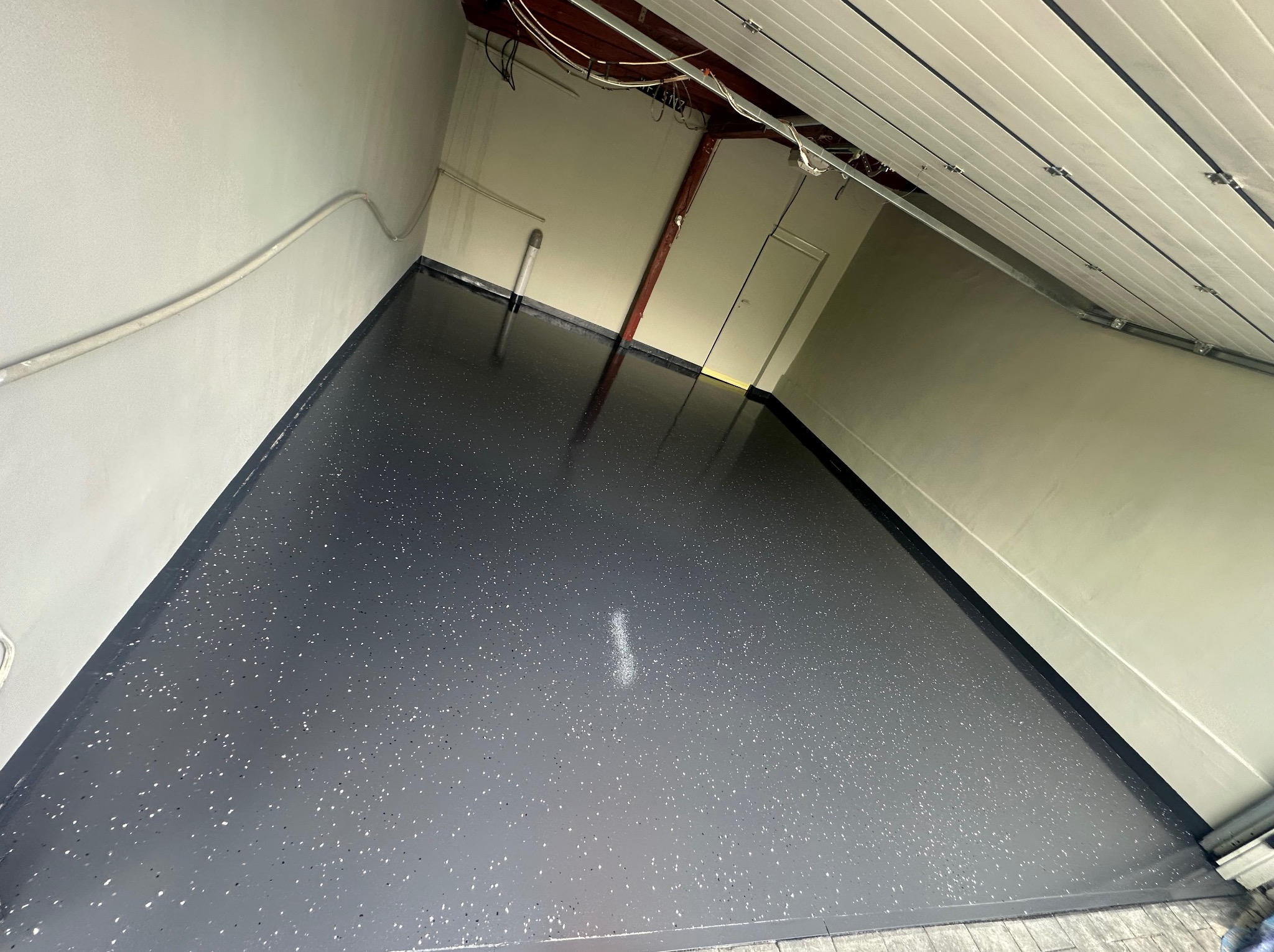 Błyszcząca, szara posadzka żywiczna z białymi i czarnymi drobinkami w garażu. Widoczne białe ściany i fragment bramy garażowej. Oświetlenie sufitowe.