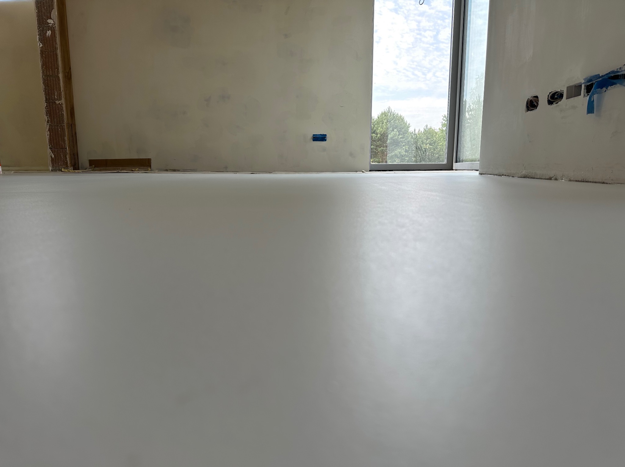 Jasnoszara, gładka posadzka żywiczna w surowym wnętrzu z oknem i widocznymi instalacjami elektrycznymi. Minimalistyczny design, naturalne światło.