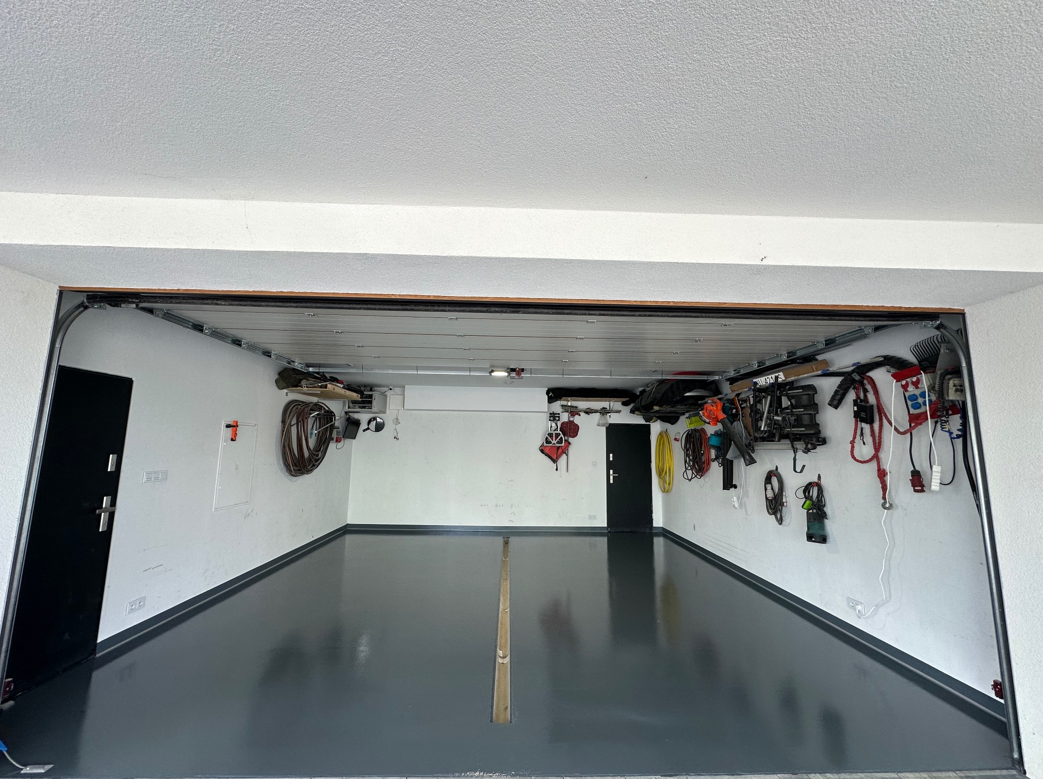 Gładka, szara posadzka żywiczna w garażu z białymi ścianami i narzędziami. Widok z zewnątrz, otwarte drzwi garażowe.