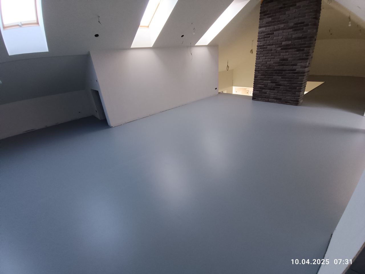 Szara posadzka żywiczna w przestronnym, jasnym poddaszu z ceglanym kominem i oknami dachowymi. Gładka powierzchnia, minimalistyczny design.