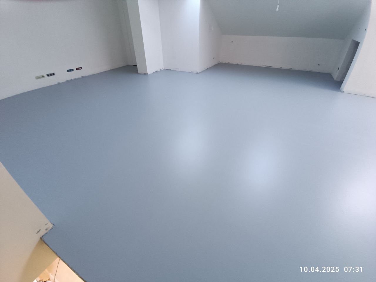 Jasnoszara, gładka posadzka żywiczna w przestronnym, jasnym pomieszczeniu z białymi ścianami i widocznymi gniazdkami elektrycznymi. Minimalistyczny design.