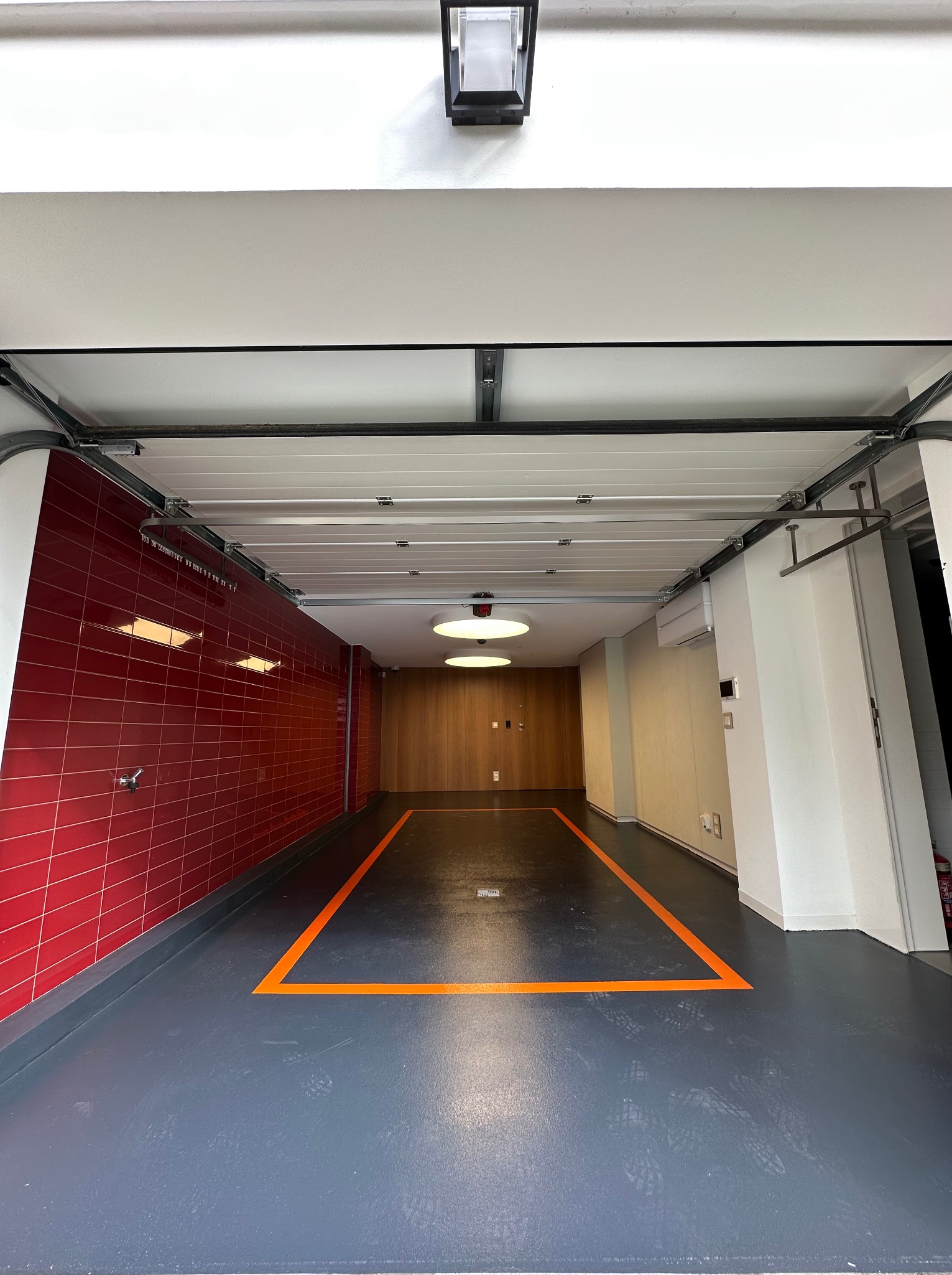 Gładka, szara posadzka żywiczna w garażu z czerwoną ścianą z płytek i pomarańczową linią wyznaczającą miejsce parkingowe. Widoczna brama garażowa i oświetlenie.