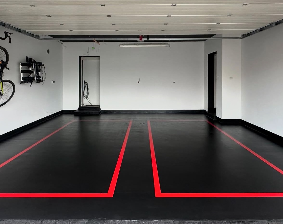 Gładka, czarna posadzka żywiczna w garażu z czerwonymi liniami wyznaczającymi miejsca parkingowe. Ściany białe, widoczny uchwyt na rower.