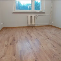 Puste pomieszczenie z nową podłogą z paneli w odcieniach brązu, białe ściany i grzejnik pod oknem z widokiem na drzewa.