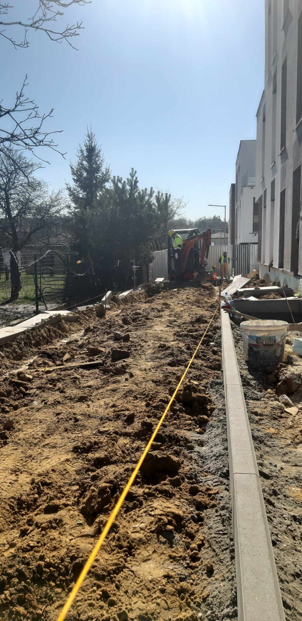 Prace ziemne przy układaniu obrzeży chodnika, w tle mini koparka i pracownicy w odblaskowych kamizelkach, widoczny sznurek wyznaczający linię, obok wiadro z zaprawą budowlaną.