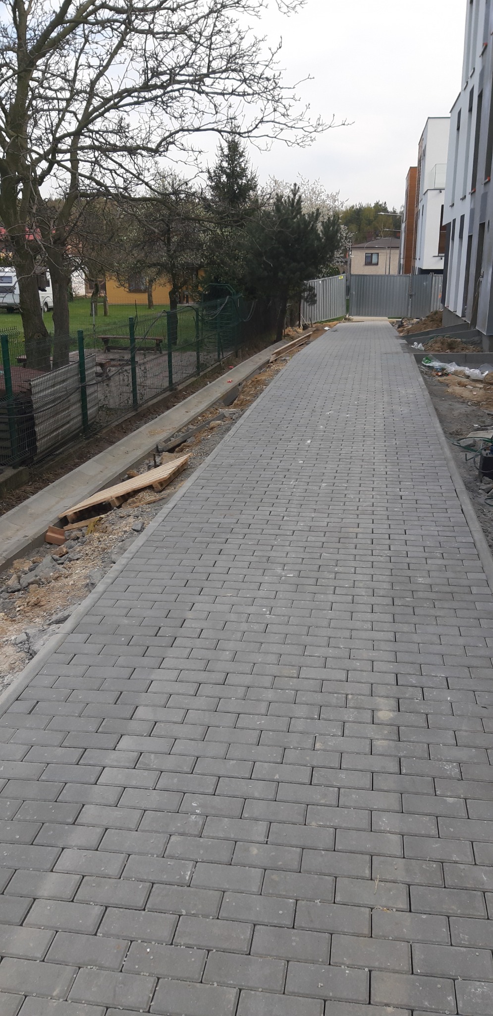 Ułożona szara kostka brukowa tworząca chodnik, widoczne krawężniki i przygotowane podłoże z boku, w tle budynek i drzewa.