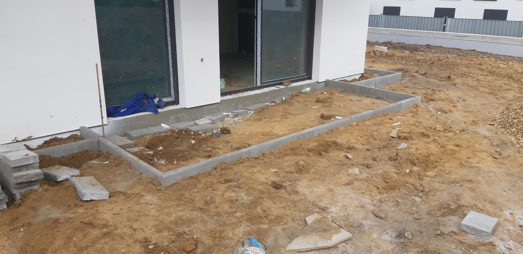 Przygotowanie terenu pod taras: ułożone obrzeża z betonowych elementów, widoczne płyty chodnikowe i piasek, budynek w tle z dużymi oknami.