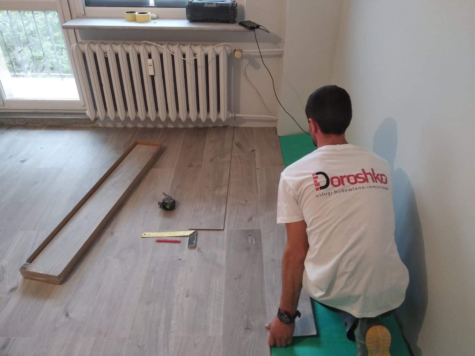 Układanie paneli podłogowych w pokoju z grzejnikiem; widoczny pracownik w koszulce z logo firmy, narzędzia pomiarowe i fragment podkładu pod panele.