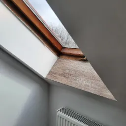 Poddasze z oknem dachowym z drewnianą ramą i parapetem imitującym drewno, widok na drzewo za oknem, szara ściana i biały grzejnik.