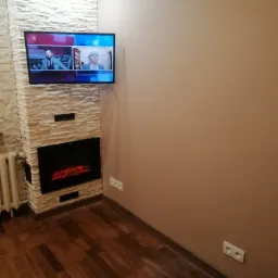 Salon z imitacją kominka obudowaną kamieniem dekoracyjnym, telewizorem na ścianie i drewnianą podłogą.